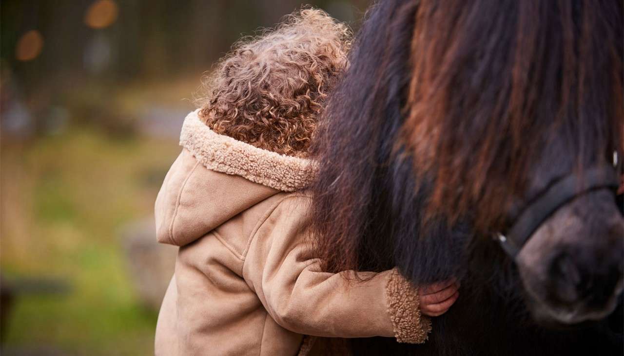 Child in tan shearling coat hugs a dark-haired pony, pressing face into its mane, one hand grasping fur. Scene outdoors with soft, blurred greenery and warm bokeh lights.