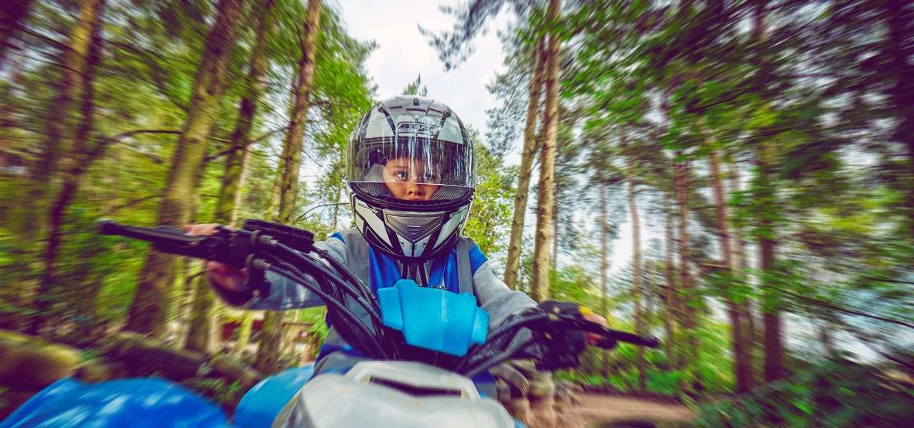 Helmeted child rides a blue ATV, gripping both handlebars, eyes focused ahead; surrounding forest blurs with motion, tall trees and a dirt path indicating speed through a wooded trail.