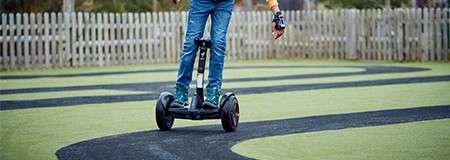 Rider steers a two-wheeled self-balancing scooter, following a winding black path on green turf, while wearing jeans and gloves; wooden picket fence and trees line the background.