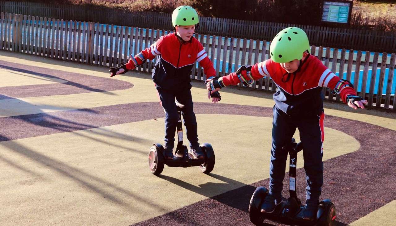 Two children ride self-balancing scooters, arms out for balance, wearing green helmets and tracksuits, on a sunlit playground with patterned surface, long shadows, and a wooden fence in the background.