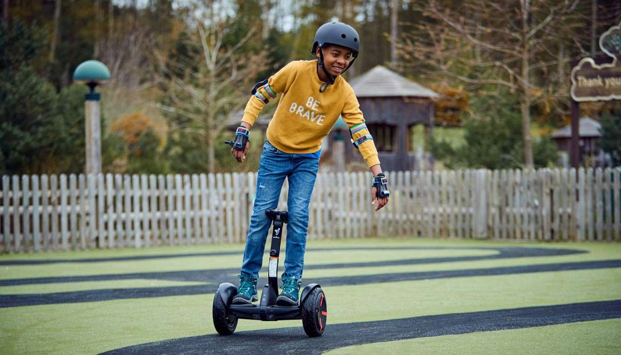 Child on a self-balancing scooter leans to turn on a winding track, wearing helmet and pads, in a park with a wooden fence and trees. Shirt text: “BE BRAVE”. Sign: “Thank you”.