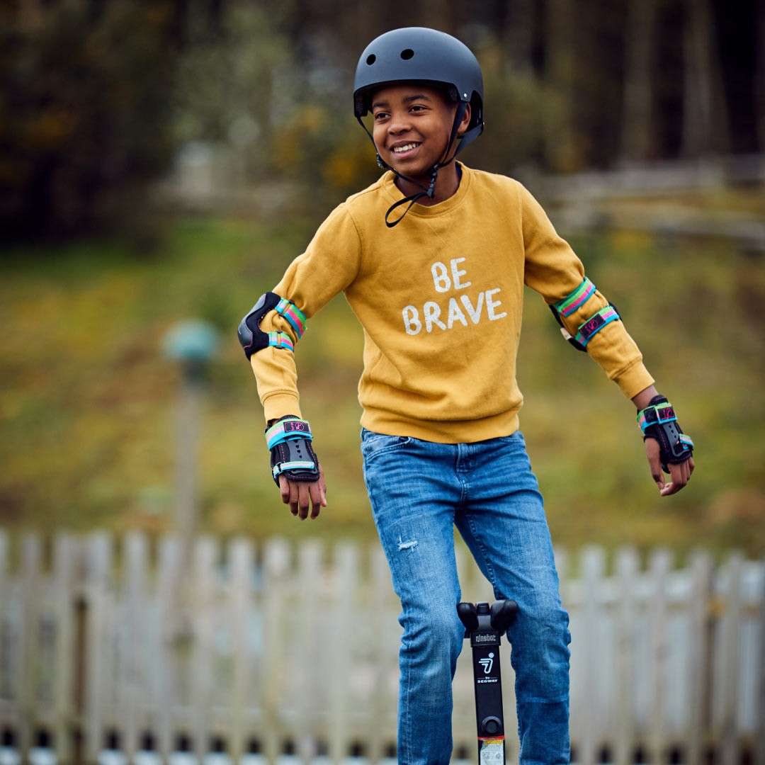 Child wearing a helmet rides a scooter, smiling. Outdoor park with fence and trees. Sweater text: “BE BRAVE”. Wrist guards show “187”. Scooter stem bears a large “J” logo.