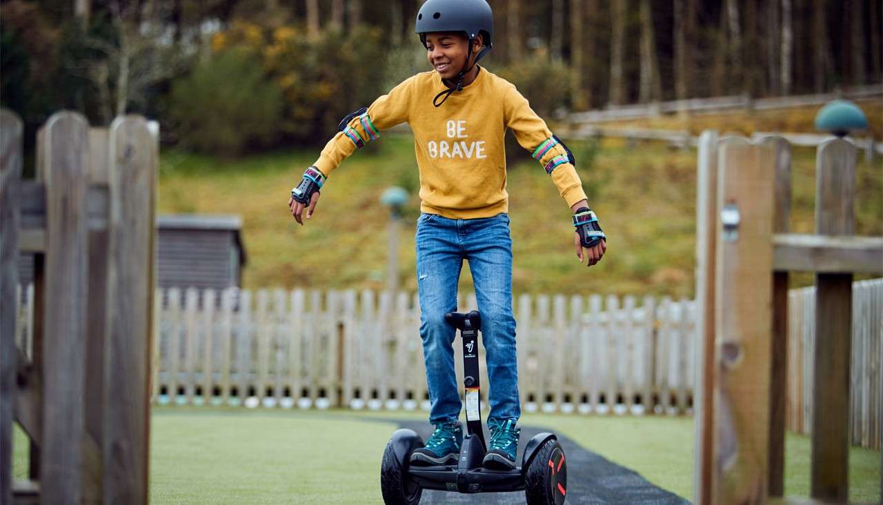 Child rides a two-wheeled self-balancing scooter, arms out for balance, wearing helmet and pads. Context: fenced outdoor area with grass and wooden structures. Text on sweatshirt: BE BRAVE.