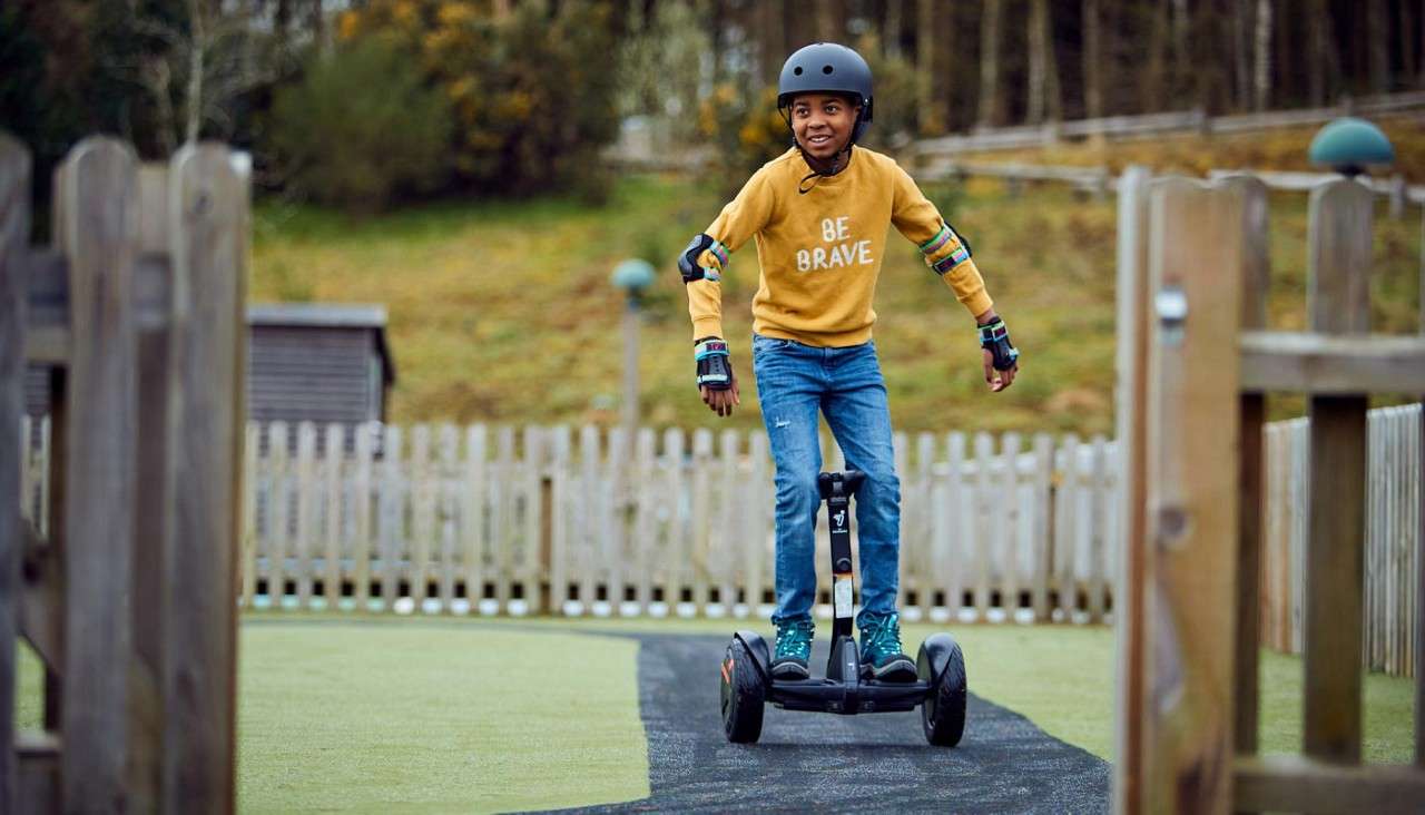 Child on a self-balancing scooter glides forward, wearing helmet and pads, along a paved path in a fenced playground. Sweater text: "BE BRAVE".