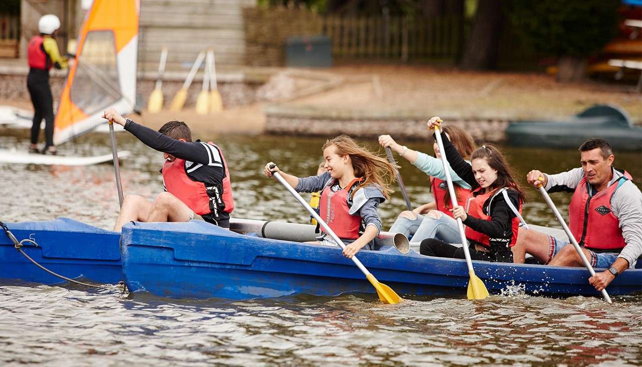 Group in a blue canoe paddles hard, water splashing, wearing red life vests. They row in sync on a calm lake, with a windsurfer and sandy shore with steps behind.