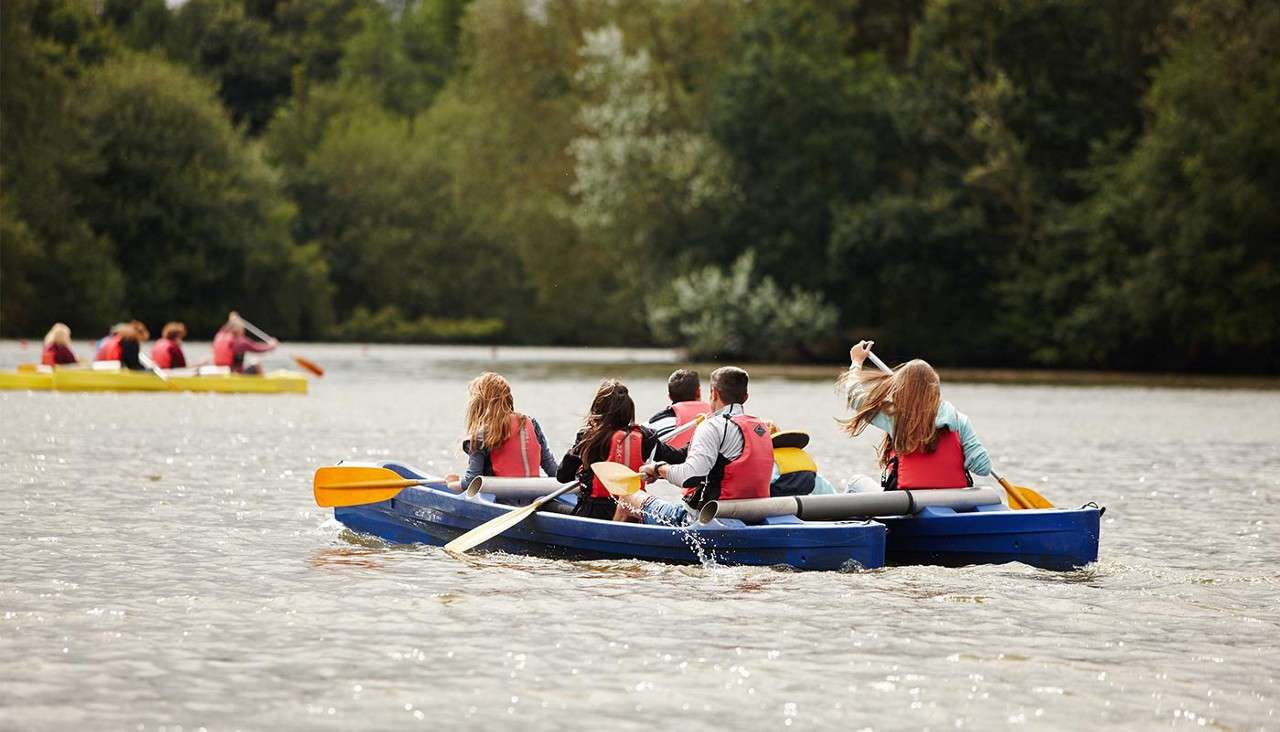 A group of people paddle a blue canoe across a calm lake, wearing red life jackets; other boats follow in the distance, with dense green trees lining the shore.