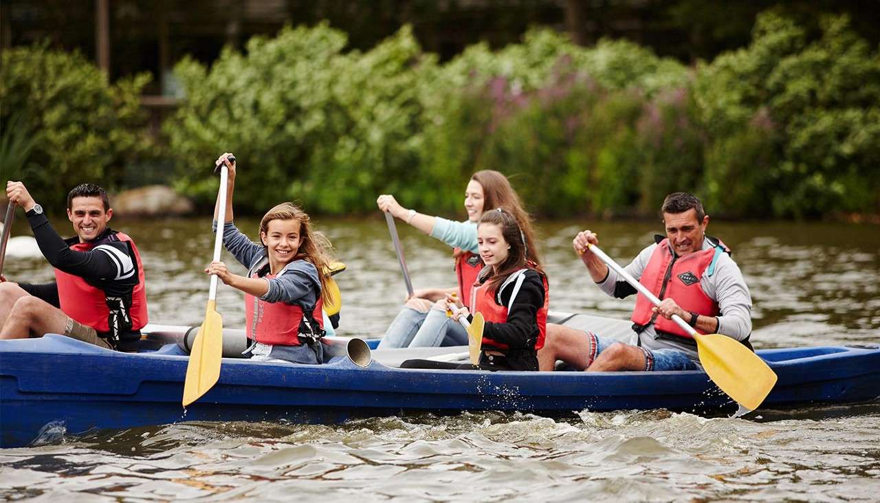 Five people in red life vests paddle a blue canoe together, smiling and splashing water, on a calm river, with green bushes and trees lining the banks in the background.