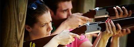 Two shooters raise shotguns to their shoulders, aiming down sights. They stand under a wooden shelter at a shooting range, concentrating, fingers near triggers, barrels pointed forward.