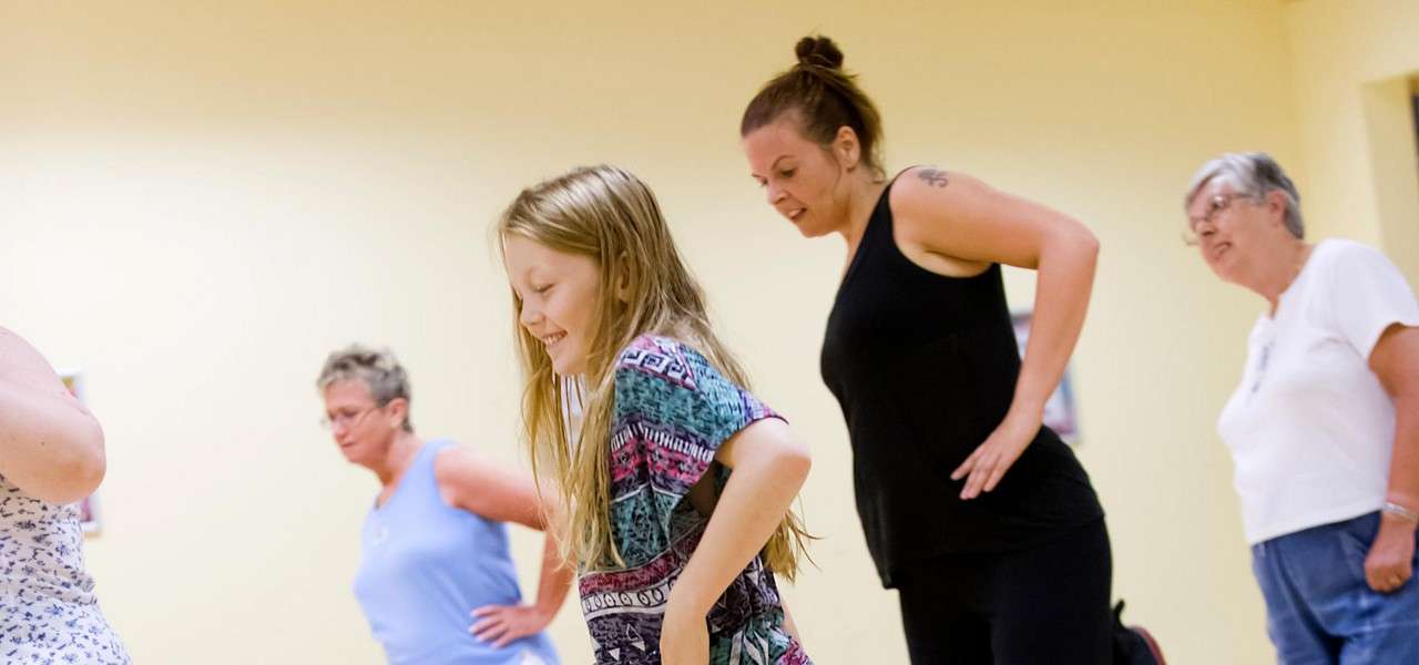 People exercise—participants bend forward with hands on hips—inside a bright studio with beige walls. A child leads in the foreground, adults follow behind, suggesting a group dance or fitness class.