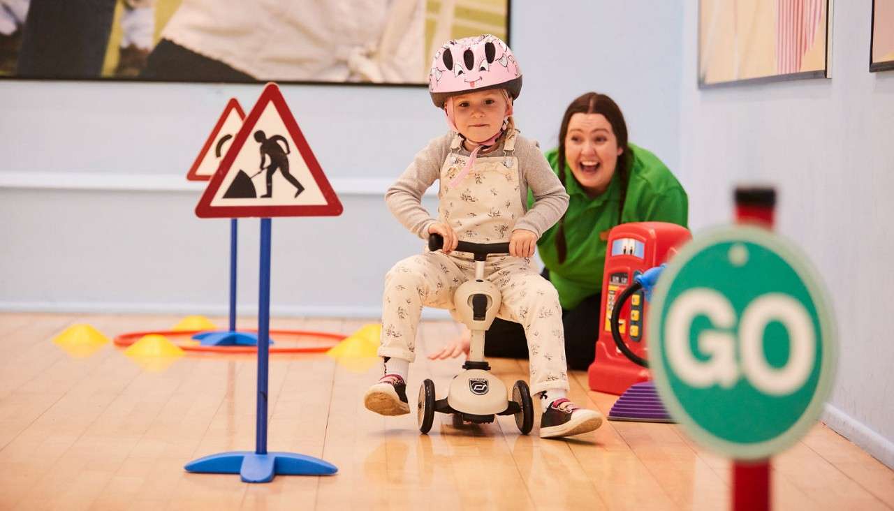 Child rides a small three-wheeled scooter, navigating cones and traffic signs, while an adult watches in an indoor play area; visible sign reads: GO.