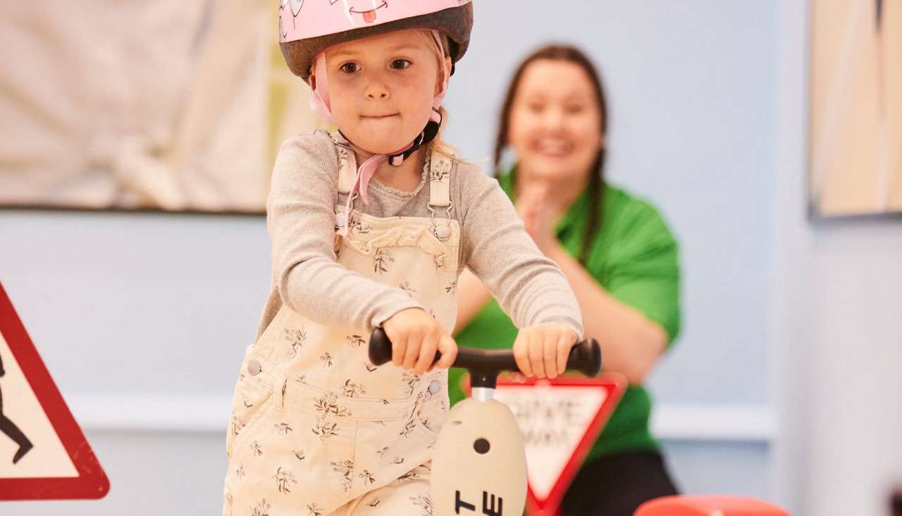 Child rides a small scooter, concentrating, wearing a pink helmet; adult in green claps encouragingly behind; toy traffic signs and pale walls indicate an indoor play or training area.