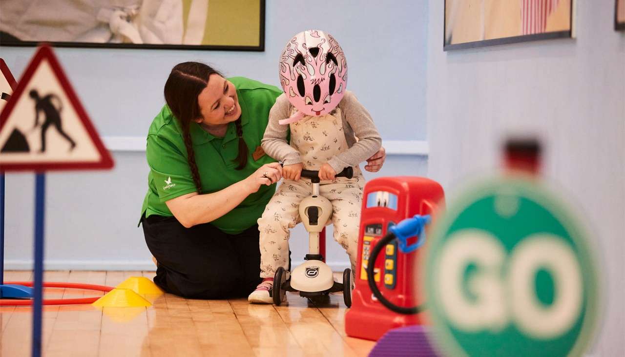 Child rides a small scooter while an adult guides them, indoors amid traffic-themed toys, cones, and signs, including a green sign reading "GO".