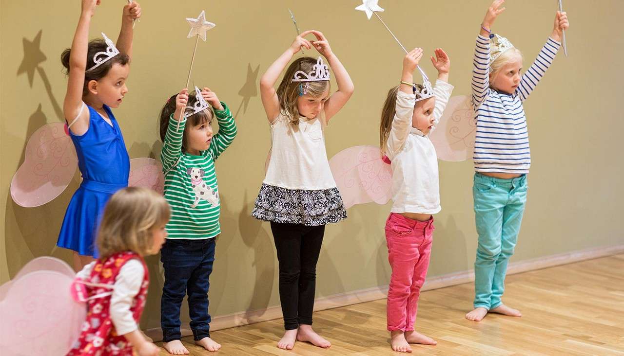 Children in fairy costumes raise star wands and arms while standing barefoot in a line, practicing a dance or play in a bright room with wooden floor and plain wall.