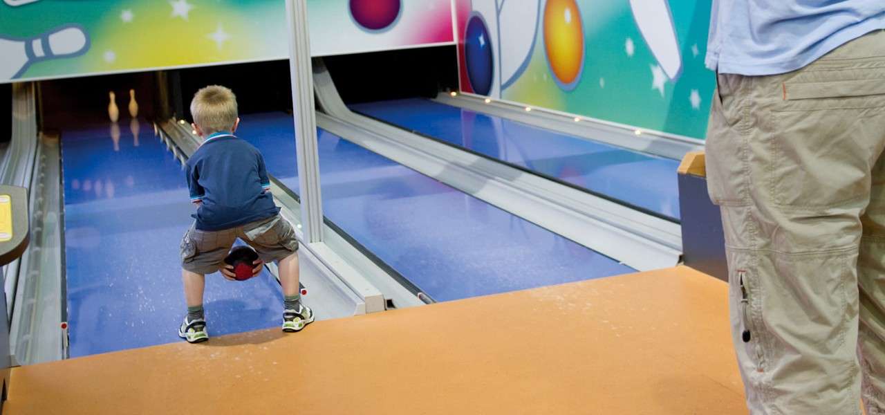 Child squats, holding a bowling ball between their legs, preparing to roll. In a bumper bowling lane, two pins stand ahead beneath a colorful mural, with an adult nearby.