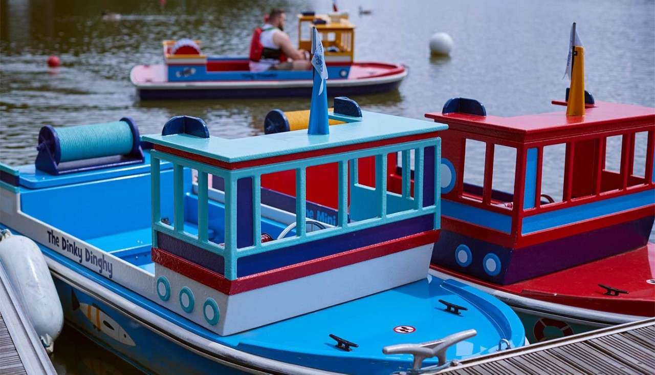Colorful miniature cabin boats are moored beside a wooden dock on a calm lake; another boat carries a person in the background. Text: The Dinky Dinghy; The Mini.