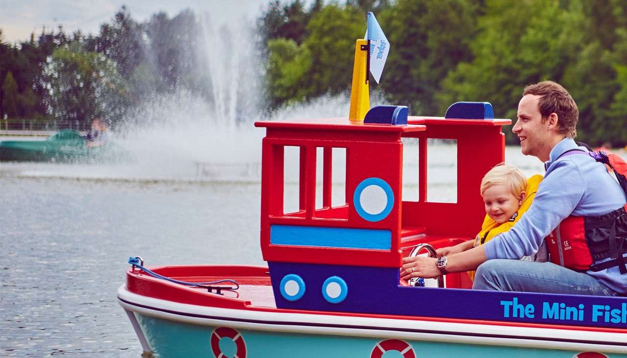 An adult and a child steer a bright red-and-blue kiddie boat labeled “The Mini Fis,” cruising on a lake with fountains, near green trees, both wearing life jackets.