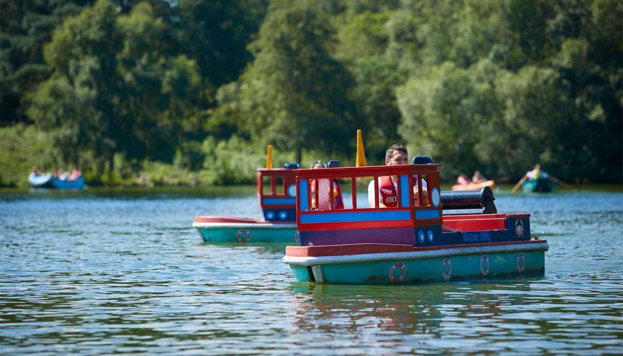 Two mini red-and-blue tugboats carry riders, cruising across a calm lake; additional boats paddle in the distance, framed by dense green trees on a bright, sunny day.