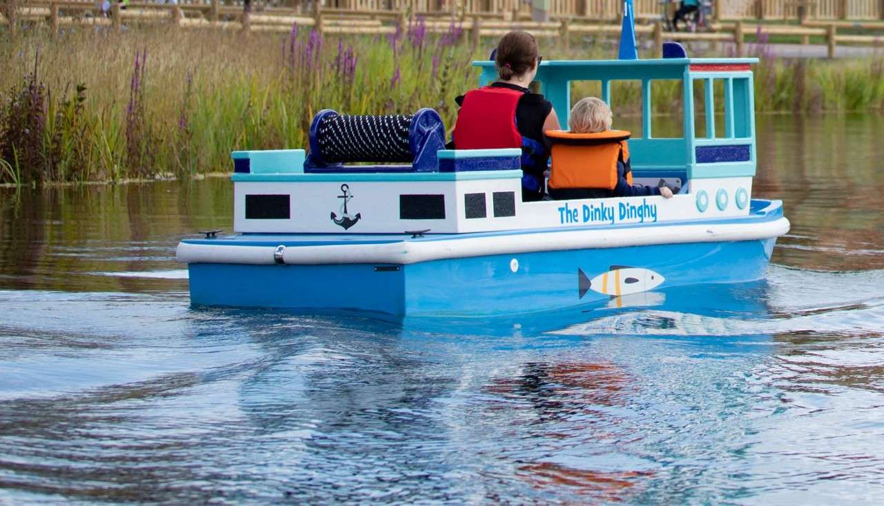 Small blue boat carries an adult and child wearing life jackets, motoring across a calm pond; reeds and fences line the shore. Visible text: The Dinky Dinghy.