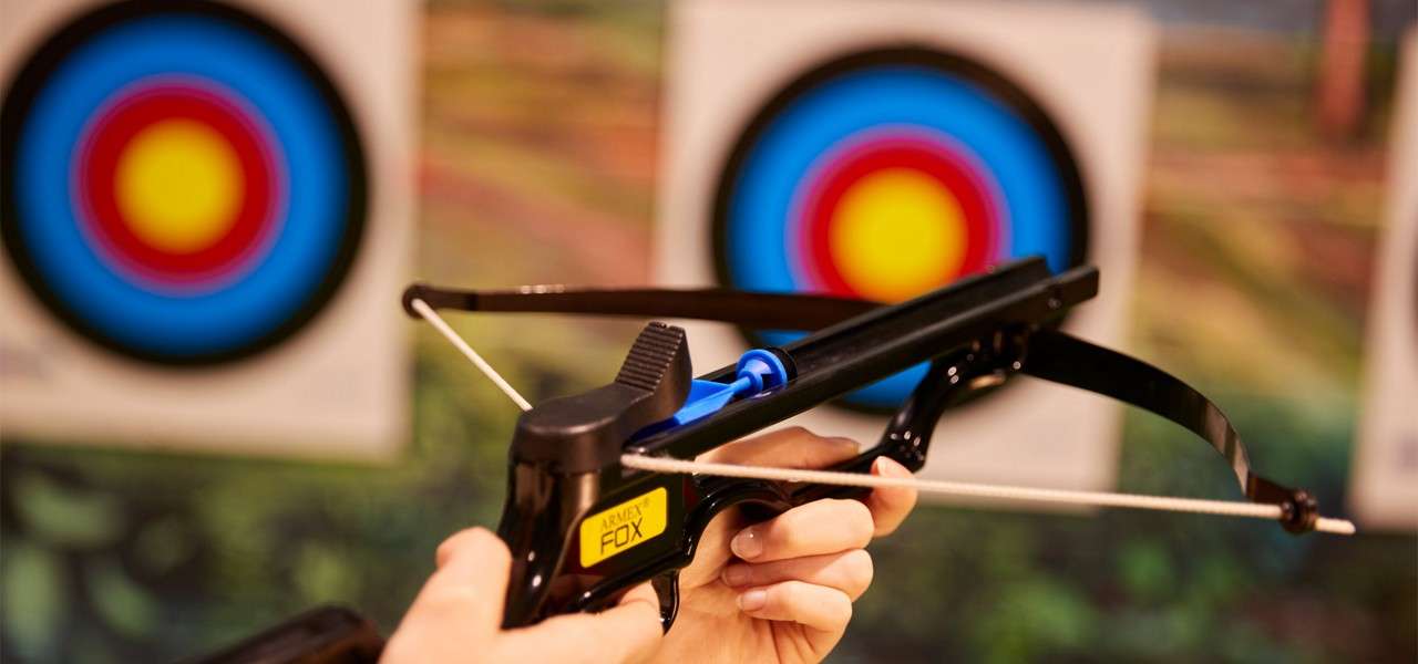 Pistol crossbow is aimed, bolt loaded, by two hands; in the background, two multicolored archery targets hang on a range wall. Text visible: ARMEX FOX.