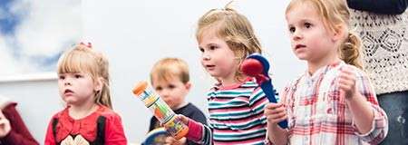 Children hold toy musical instruments and watch intently, standing side-by-side. In a bright indoor classroom, another child and an adult partially visible in the background observe.