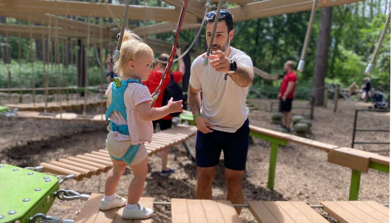 Toddler in a blue safety harness steps onto a suspended wooden bridge while an adult guides and points forward; ropes course set in a wooded park with other participants nearby.