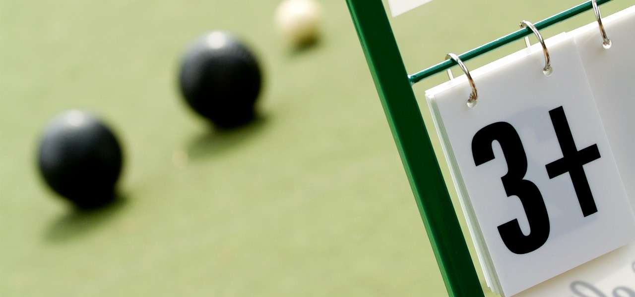 Scoreboard card displays "3+", hanging on a green frame, while two black bowls and a smaller white jack sit blurred on a lawn bowling green.