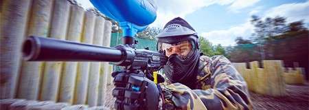 Paintball player aims a marker with hopper and barrel, advancing forward; wearing camouflage and protective mask; outdoor field with wooden bunkers and trees, motion blur emphasizing fast movement.