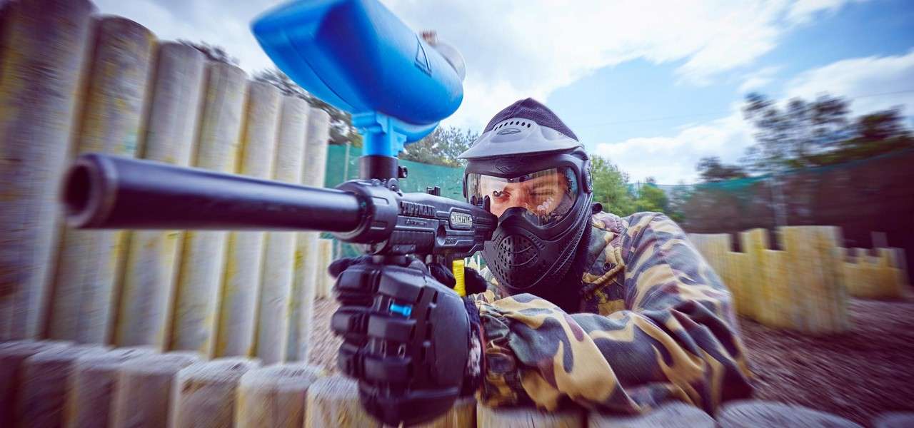 Paintball player aims a Tippmann marker while crouching behind stacked wooden logs on an outdoor field, wearing a protective mask and camo jacket; background trees and netting blur with motion. Text: TIPPMANN.