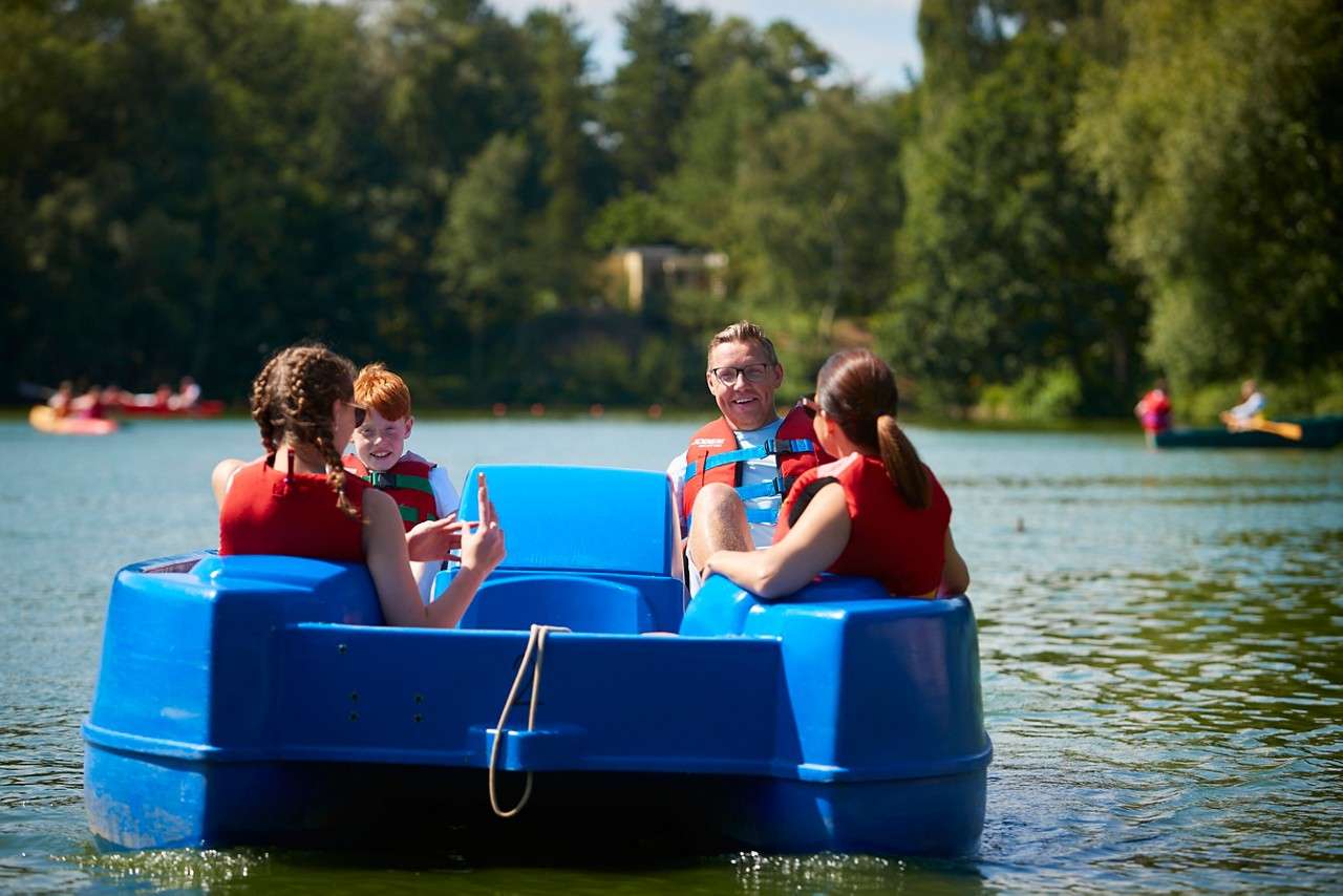 Green pedal boat carries four people, moving across a calm lake; nearby sailboats glide and trees line the shore. Text on boat reads: "pioner tamar".