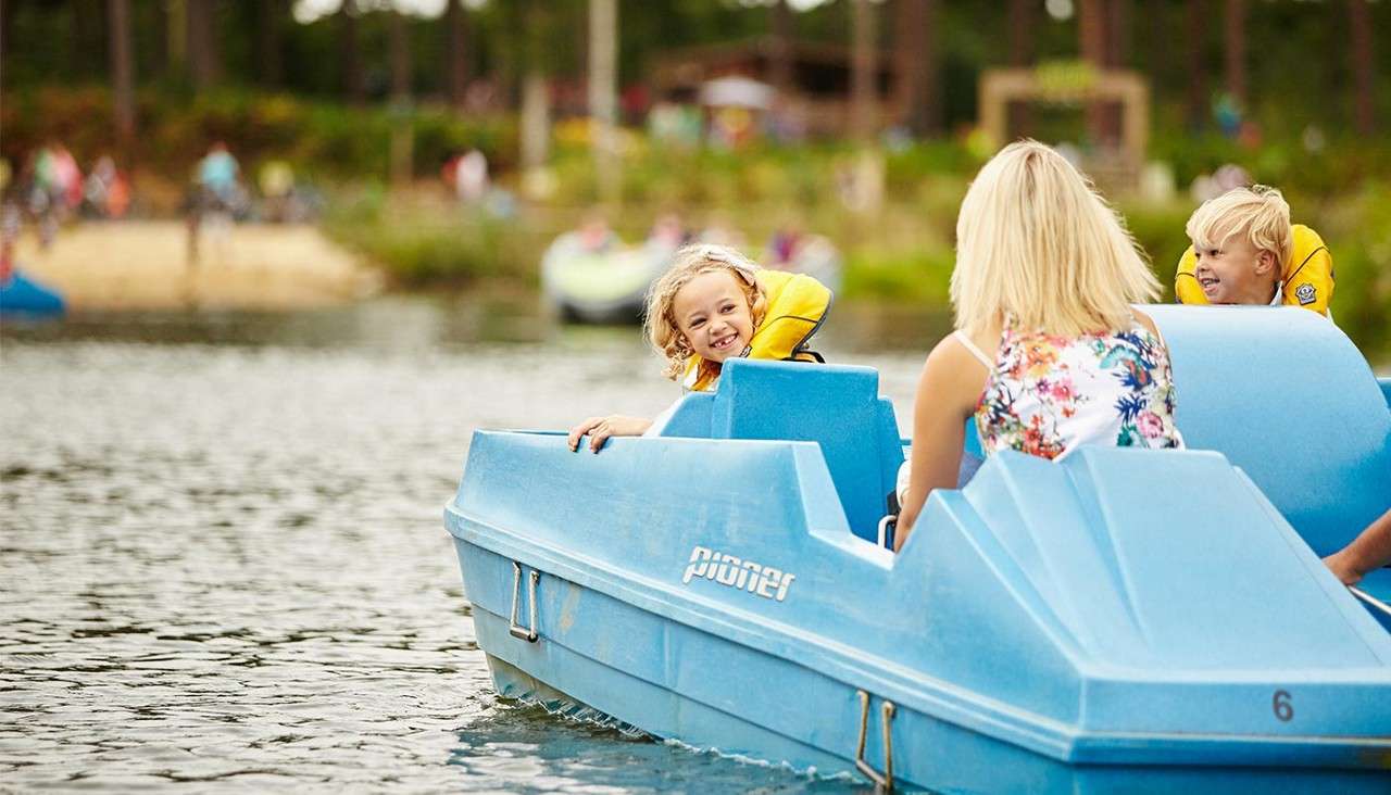 Blue pedal boat carries smiling children and an adult, gliding across a calm lake; forested shore with people and a sandy beach in the background. Text: pioner, 6.