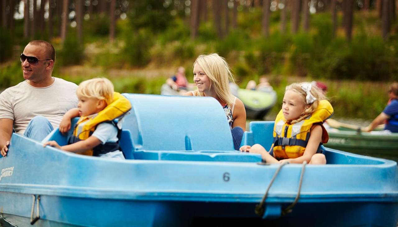 Family in a blue pedal boat glides, children wearing yellow life jackets while adults smile. Around them, other boats share a lake beside a forested shoreline. Text: 6.