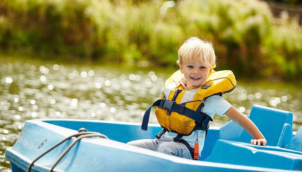 Child in a yellow life jacket smiles while sitting in a blue paddle boat, holding the side, on a sunlit lake with green shore in background. Text: 6.