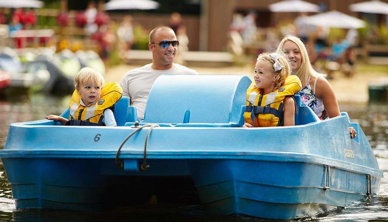 Blue pedal boat carries two children and two adults wearing yellow life jackets, cruising calmly on a small lake; background shows blurred shoreline with people and umbrellas. Text: 6.