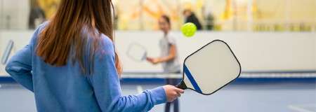 Pickleball player grips a square paddle, preparing to volley a bright ball; indoors, a net divides the court while another player waits in the blurred background.