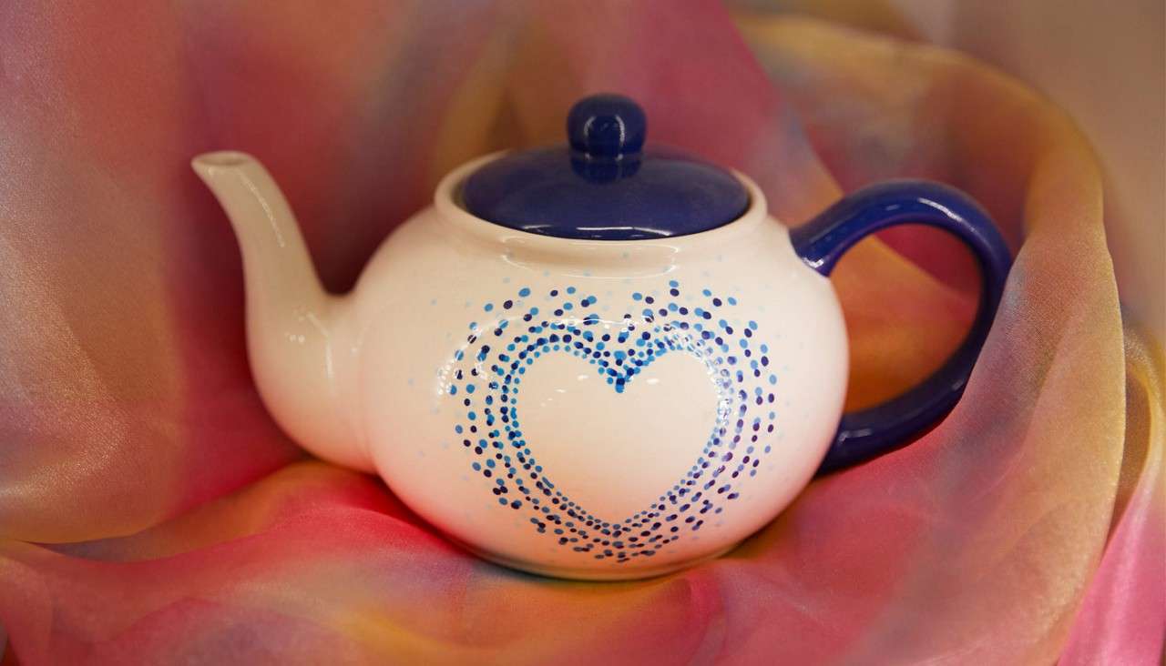 White ceramic teapot rests on draped, multicolored fabric; blue lid and handle. Dotted blue heart pattern decorates the side, while soft lighting highlights glossy surfaces and gentle folds.
