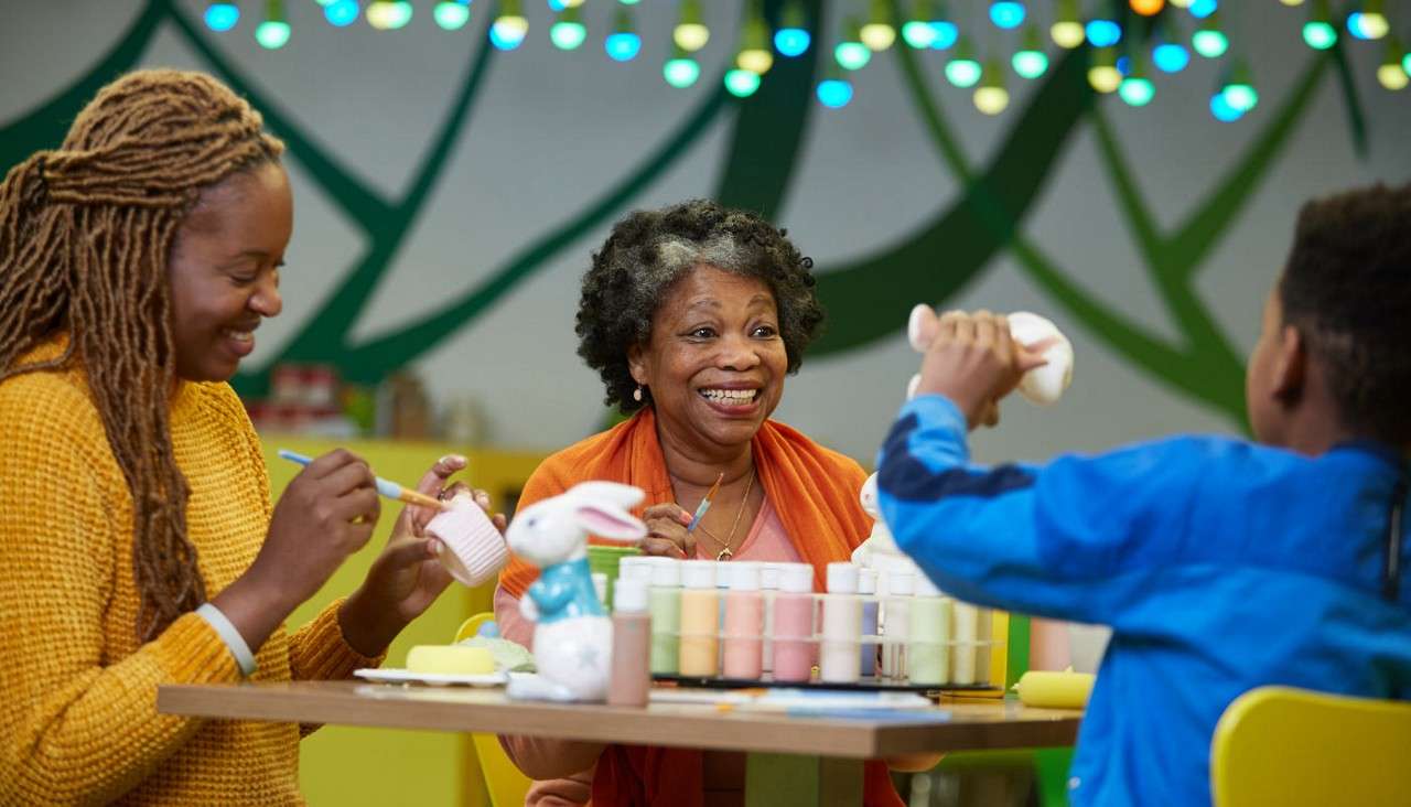 Three people paint ceramic pieces at a table, smiling. They hold brushes and mugs beside rows of paint bottles and a ceramic rabbit, under colorful lights in a studio.