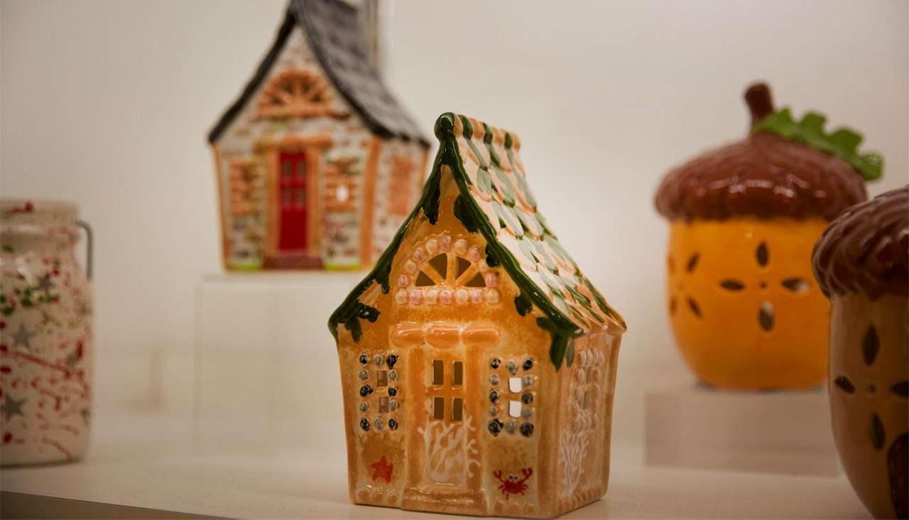 Ceramic house figurine sits on a white display, showing cutout windows and green-tiled roof; behind, a blurred ceramic cottage, acorn-shaped containers, and a jar complete the shelf arrangement.