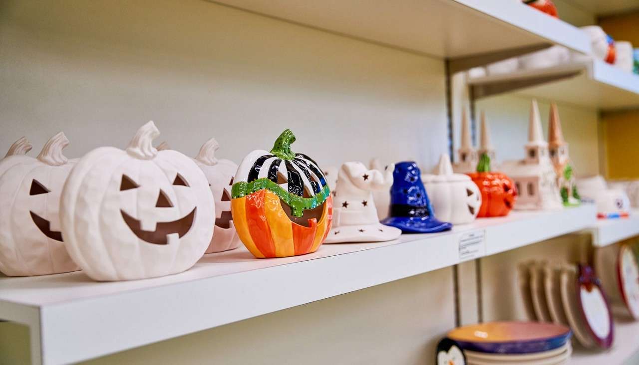 Ceramic jack-o’-lanterns sit displayed on a white shelf, some unpainted, one brightly painted; nearby witch-hat figurines and castle decorations line the shelves in a craft or holiday shop.