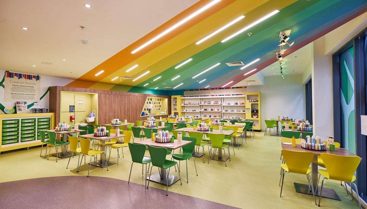 Art tables display paint bottles, awaiting use, in a bright studio. Rainbow ceiling, green-and-yellow chairs, shelves of ceramics, and supply drawers surround. Visible text: "Creation Station"; other small text unreadable.