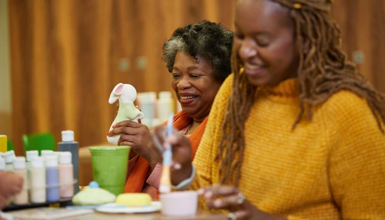 Two people paint ceramics at a craft table in a studio, one smiling while holding a mouse figurine; paints, brushes, cups, and supplies are scattered around against a wooden background.