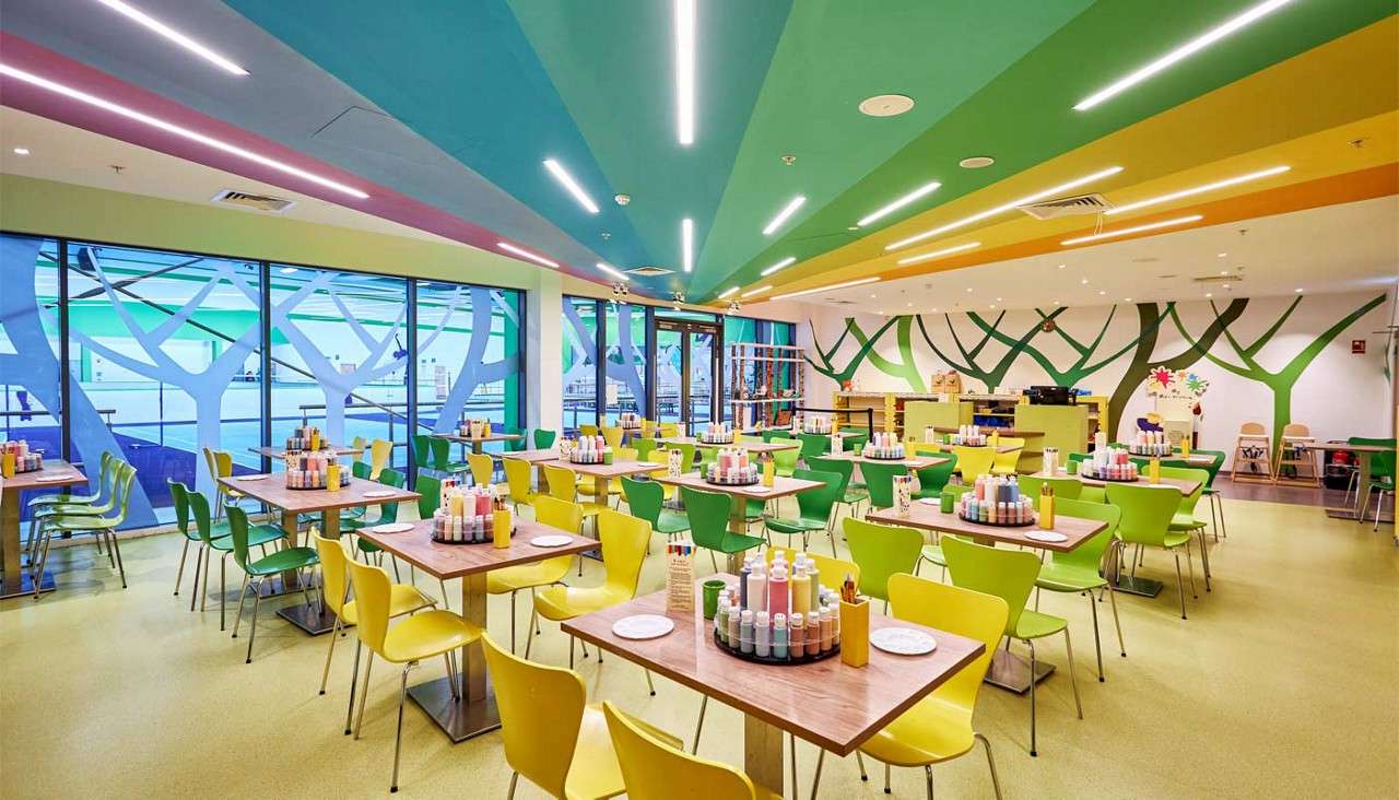 Tables with paint bottles and plates wait neatly arranged for crafters in a bright studio with yellow-green chairs, striped ceiling lights, tree murals, and large window panels.