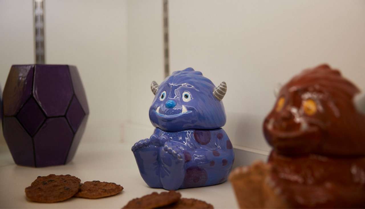 Blue ceramic monster cookie jar sits smiling with feet forward, flanked by cookies. On a white shelf, a faceted purple container and a blurred brown monster jar frame the scene.