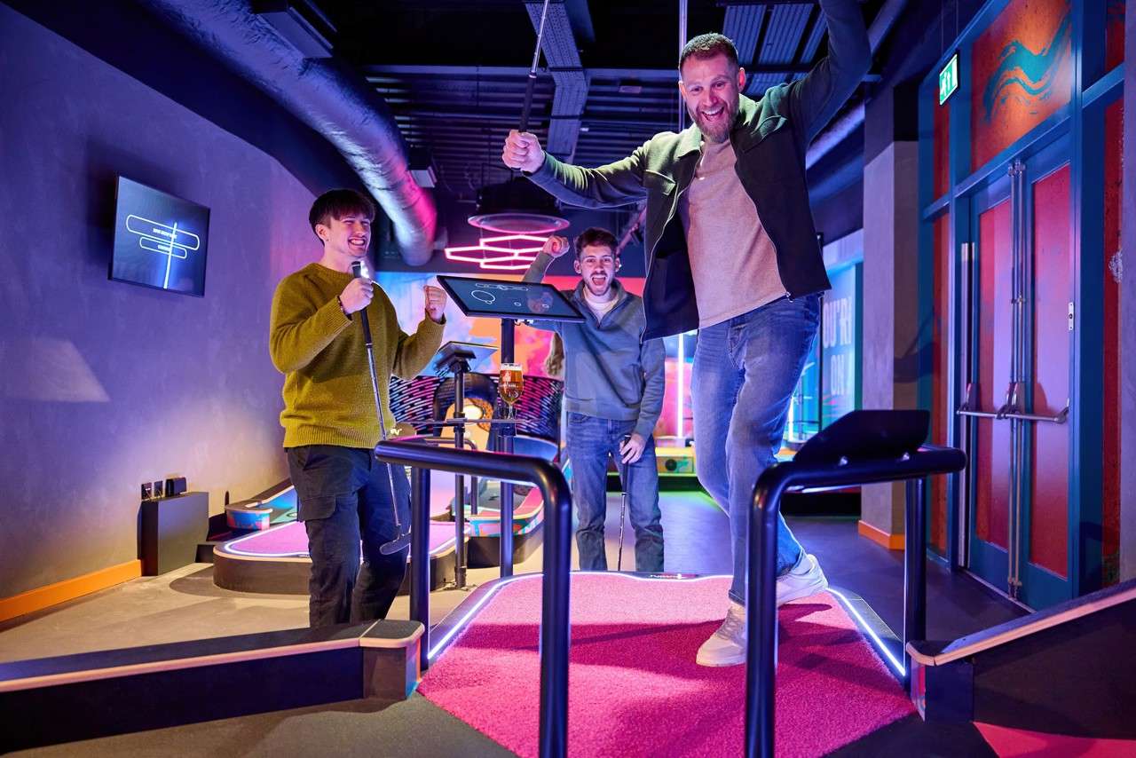 Three adults play mini-golf, cheering as one steps onto a raised pink putting platform; others hold putters nearby. Neon lights, digital score screens, and a beer glass surround them. Text: EXIT.