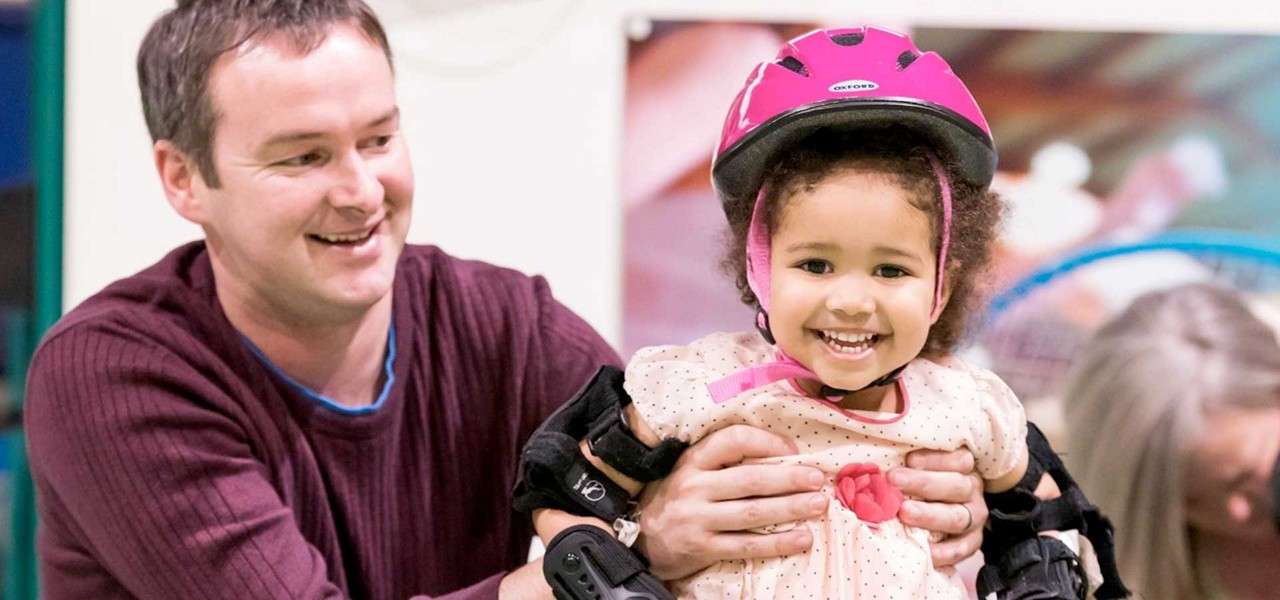 Child smiles, wearing a pink helmet and protective pads, while an adult holds her sides; indoors, another adult nearby and a blurry, colorful background suggests a play or training area.