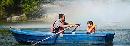 Rowboat carries an adult rowing and a child wearing an orange life jacket. On a calm lake near a spraying fountain, with green trees and rocks along the shore.