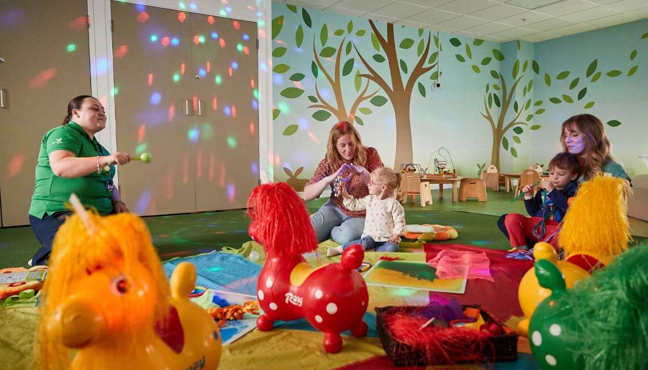 Children and adults play, shaking instruments and bouncing inflatable horse toys, in a colorful playroom with tree murals, mats, and disco-like lights. Visible text: Rody, Happy.