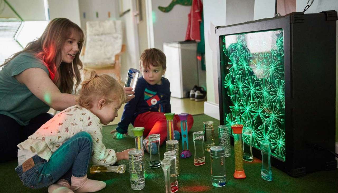 Two children play, stacking and examining sensory liquid timers, while a caregiver supports them; context: carpeted indoor playroom beside a glowing green fiber-optic light panel.