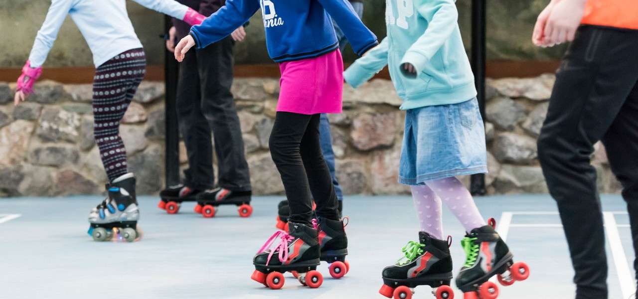 Children glide on quad roller skates, arms out for balance, on a smooth rink beside a stone wall. Visible text: "80", "AP".