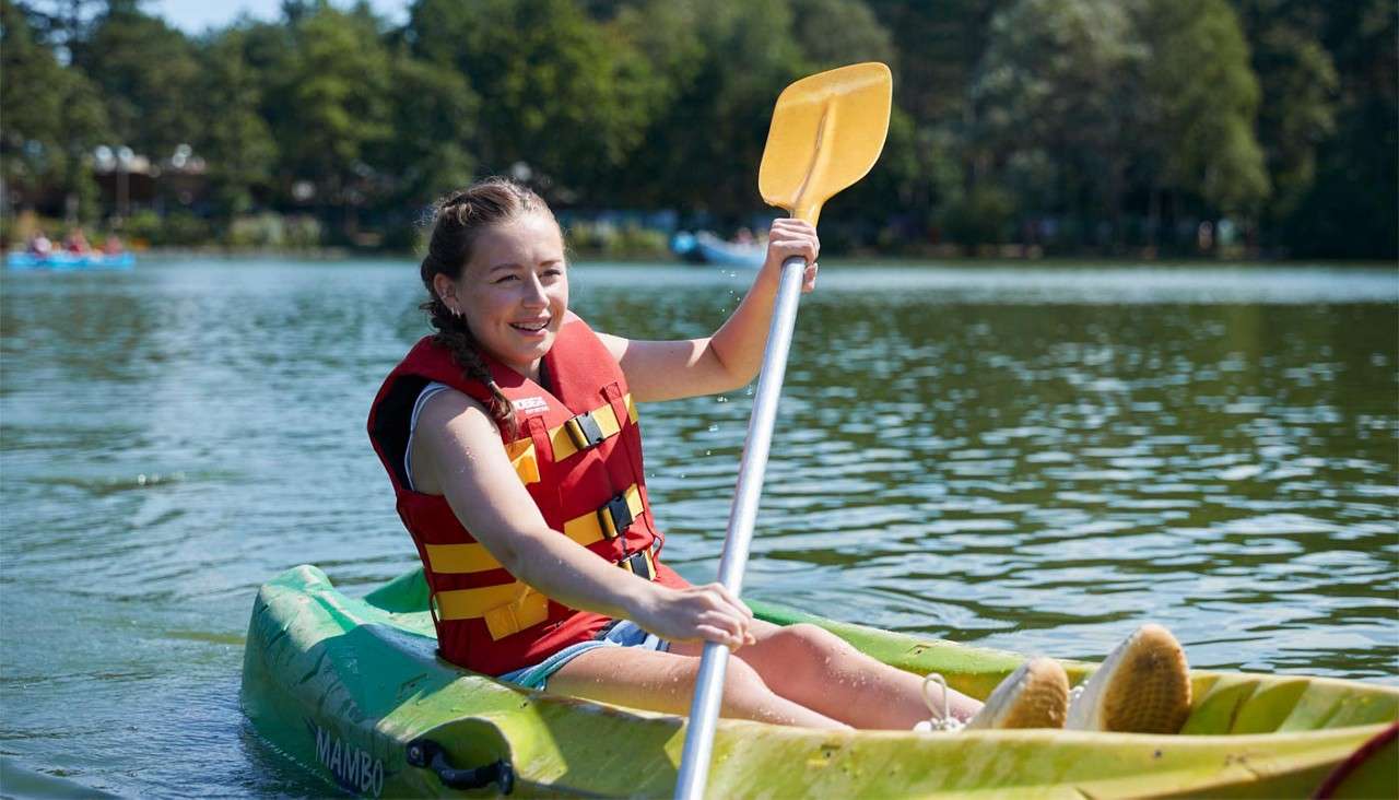 Person in a red life vest paddles a green kayak; context: calm lake with surrounding trees and distant kayaks. Text: MAMBO.