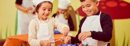 Two children in aprons and a chef hat mix ingredients with sticks in bowls, smiling; in the background, another child in chef attire works at table in decorated classroom.